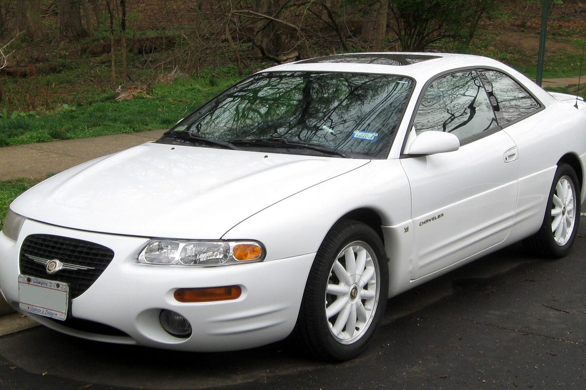 A white Chrysler Sebring two-door coupe, front left view, parked on wet pavement beside a grassy slope