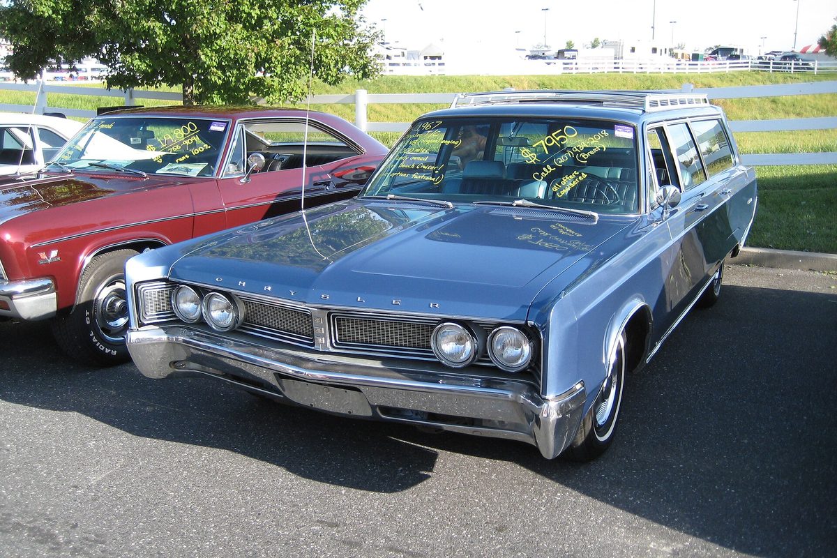 A blue 1967 Chrysler Town and Country station wagon with a roof rack and sale notes written on its windshield, parked next to a red car in the car corral at the Antique Automobile Club of America's fall meet in Hershey, Pennsylvania