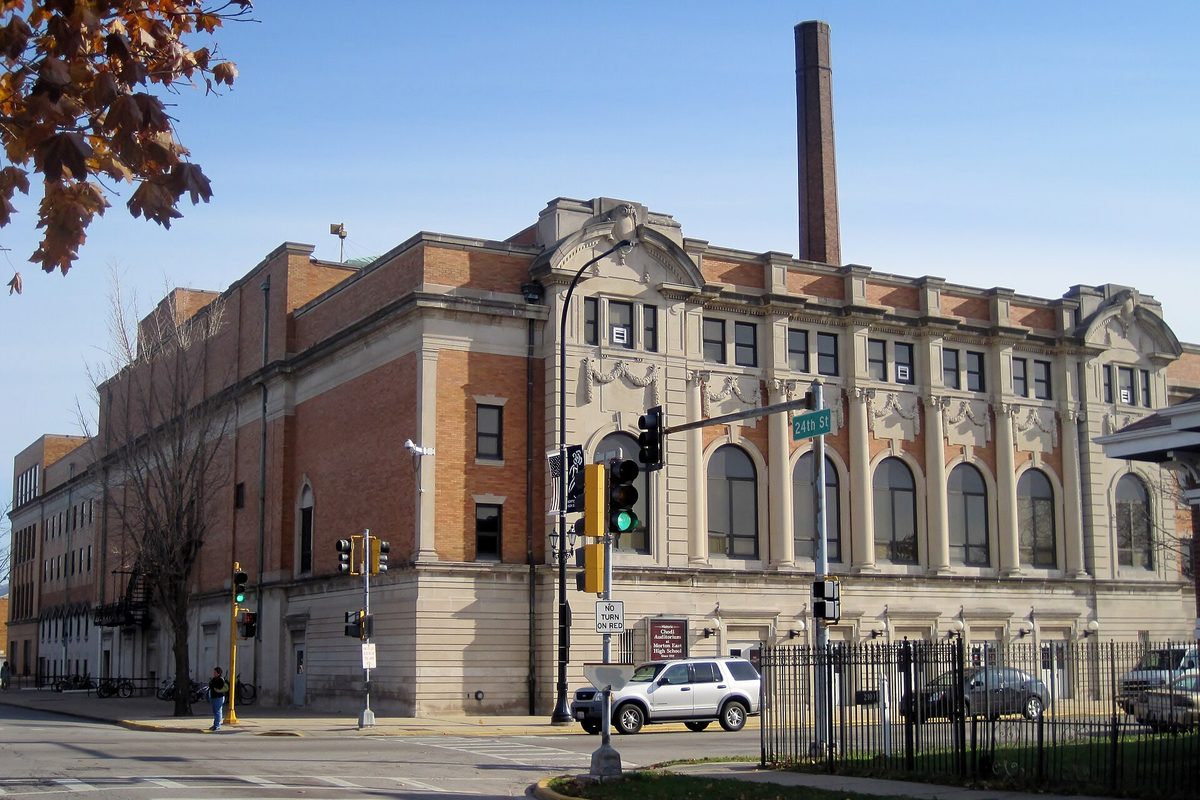 The brick and carved stone auditorium of J. Sterling Morton High School East in Cicero, with a tall smokestack, at a corner with traffic lights and a 24th St sign