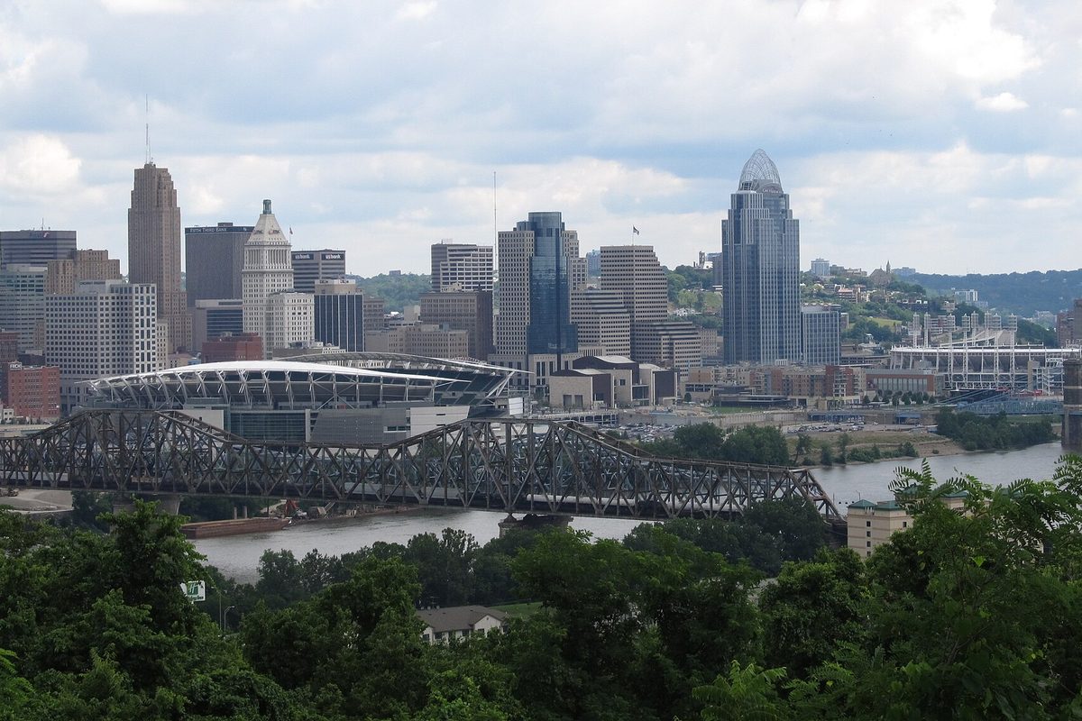 Downtown Cincinnati's towers and the riverfront football stadium behind a steel truss bridge over the Ohio River, seen through treetops from Devou Park