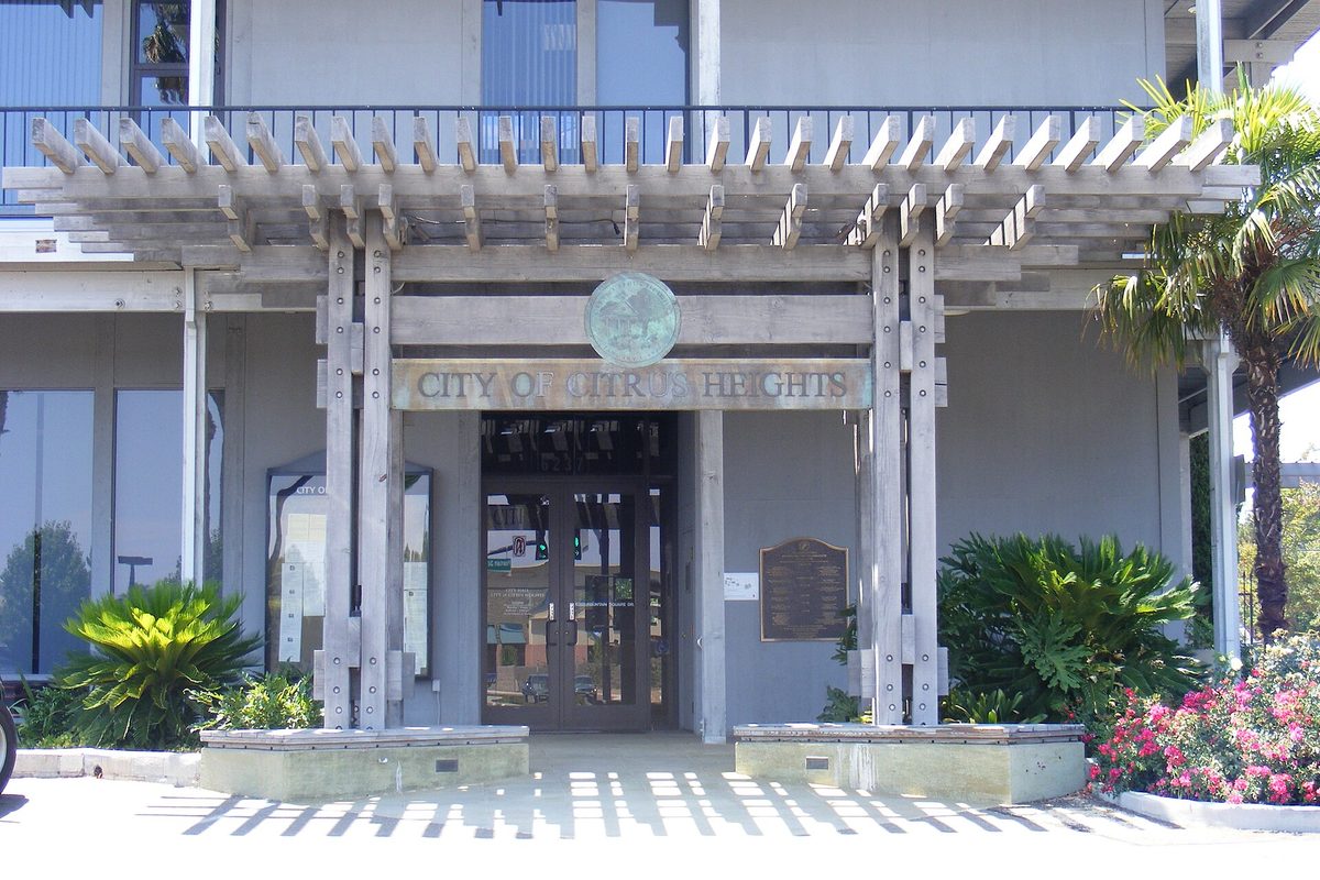 The entrance to Citrus Heights City Hall, a wooden pergola over glass doors with the city seal, framed by palms, shrubs and pink flowers