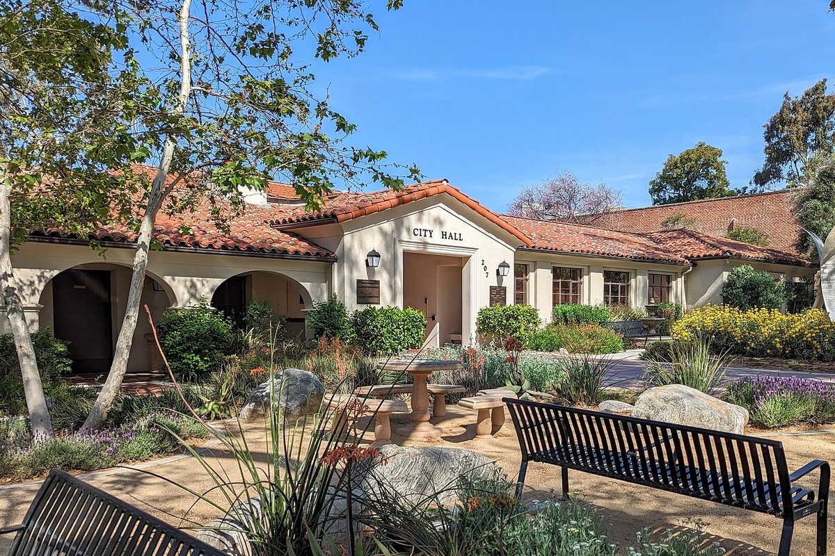 Claremont City Hall, a white Spanish-style building with a red tile roof and arched porch, behind a garden of flowering plants, boulders and black benches