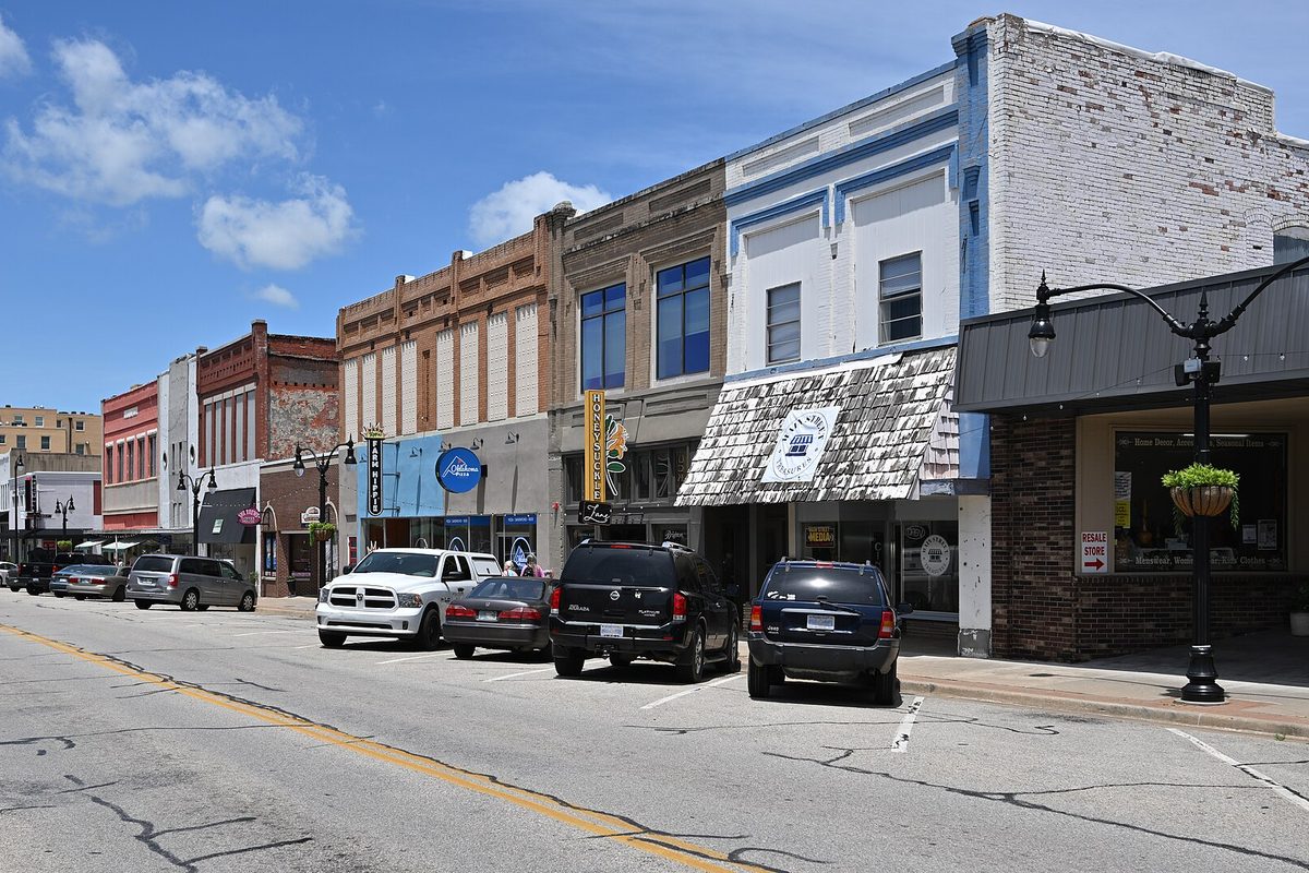 A row of brick and painted storefronts in downtown Claremore on Will Rogers Boulevard, with pickups and SUVs parked at the curb under a blue sky