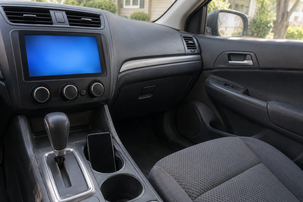 A car dashboard with a blank blue infotainment screen, three climate knobs, an automatic gear lever and a phone resting in the cupholder