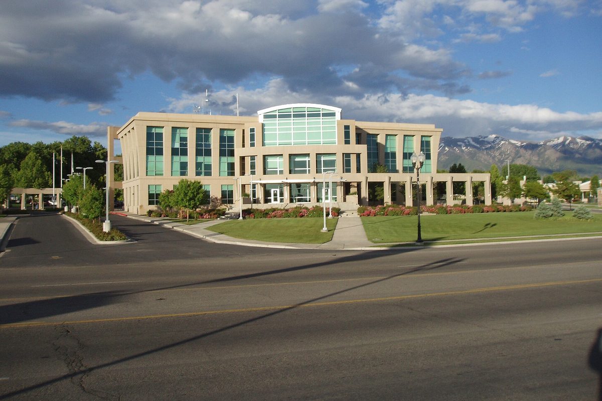 Clearfield's three-story tan Municipal and Justice Center with a glass-fronted middle section, a lawn and young trees, and snow-capped mountains behind it