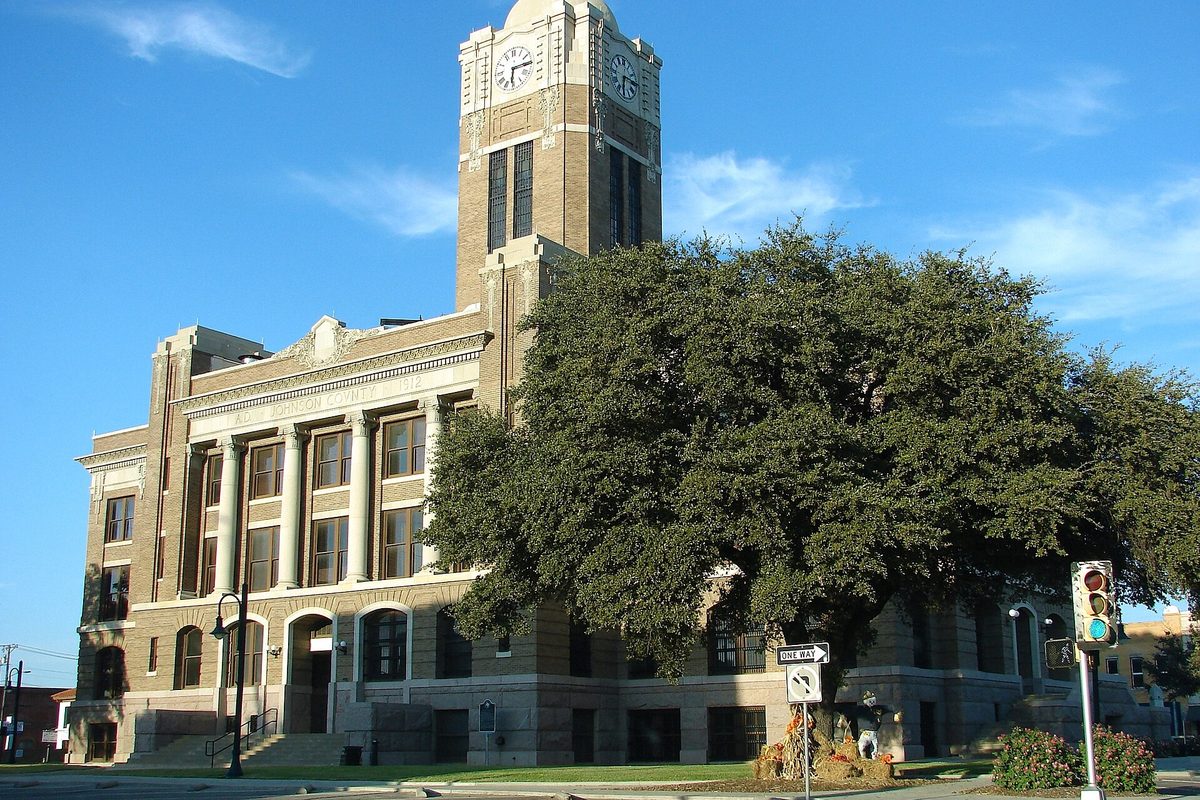 The Johnson County Courthouse in Cleburne, a tan stone building with columns and a tall clock tower, beside a broad live oak under a blue sky