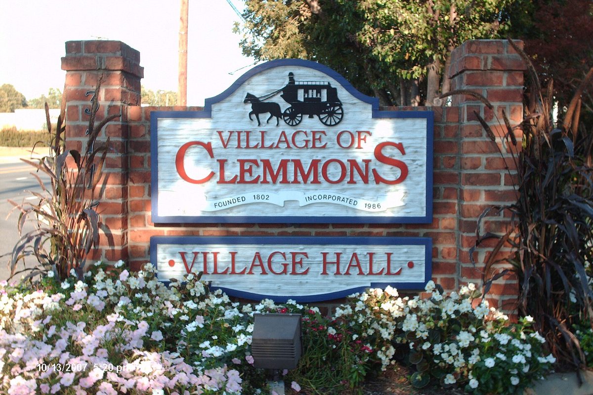 A white wooden sign reading Village of Clemmons, Village Hall, with a horse-drawn coach on top, set between brick pillars above beds of white and pink flowers
