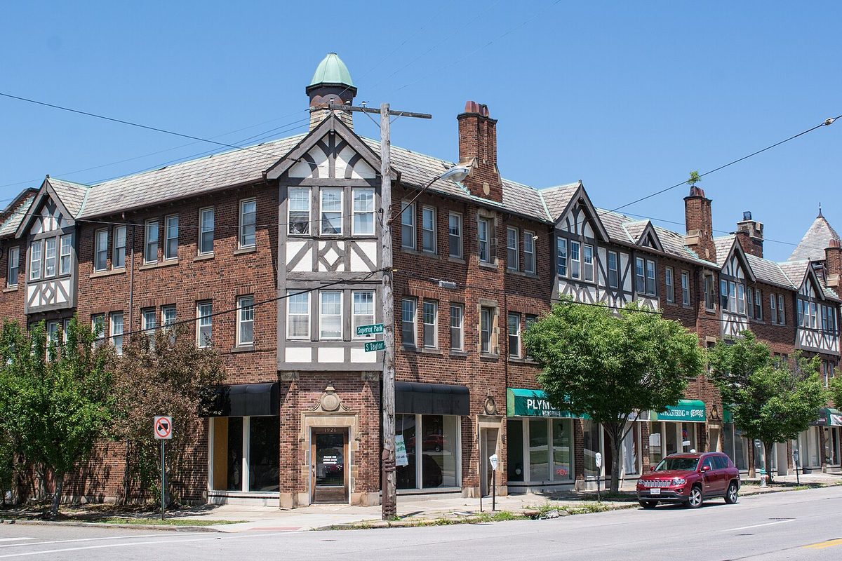 A three-story Tudor Revival brick building with half-timbered gables and shops at street level on South Taylor Road in Cleveland Heights, a red SUV parked at the curb
