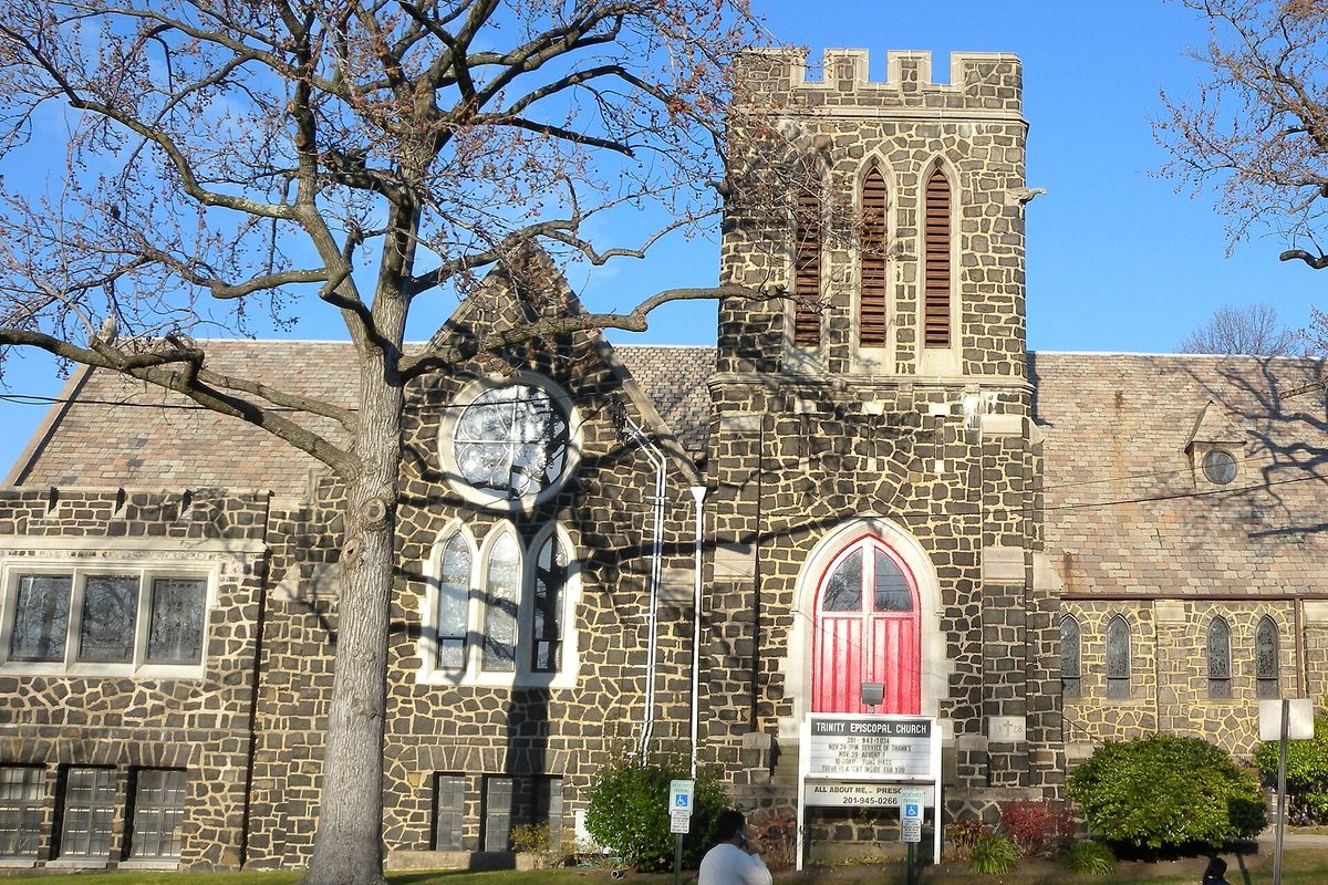 Trinity Episcopal Church in Cliffside Park, a dark fieldstone church with a square crenellated tower and a red arched door, beside a bare tree on a sunny day