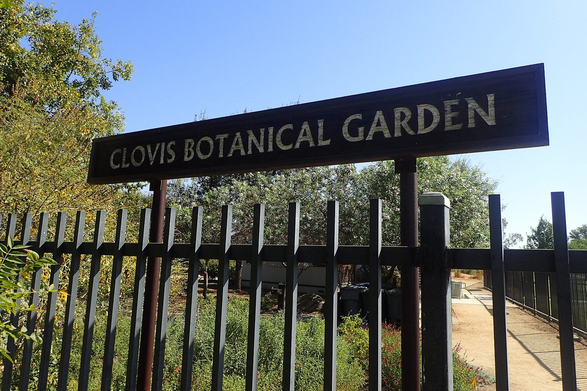 A dark sign reading Clovis Botanical Garden on posts above a black iron fence, with trees and flowering plants behind it under a clear blue sky