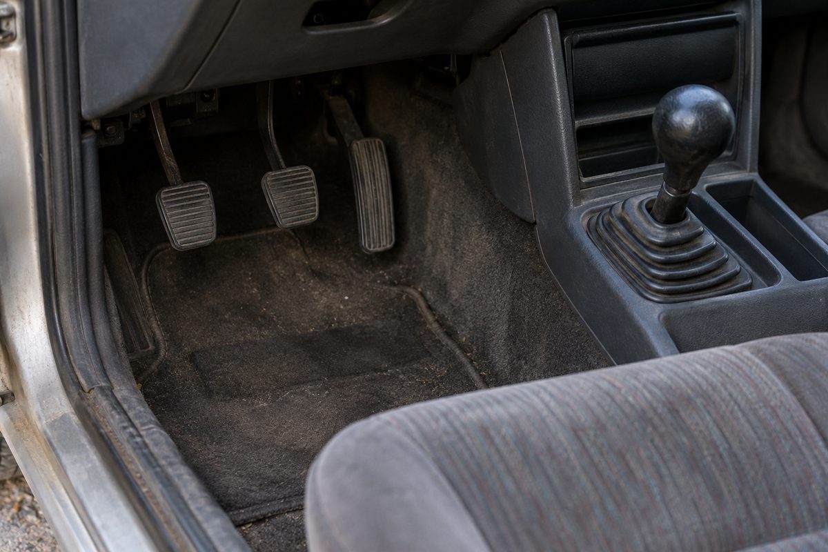 The driver's footwell of an older manual car, showing three worn pedals, dark carpet and a black gear shifter beside the seat
