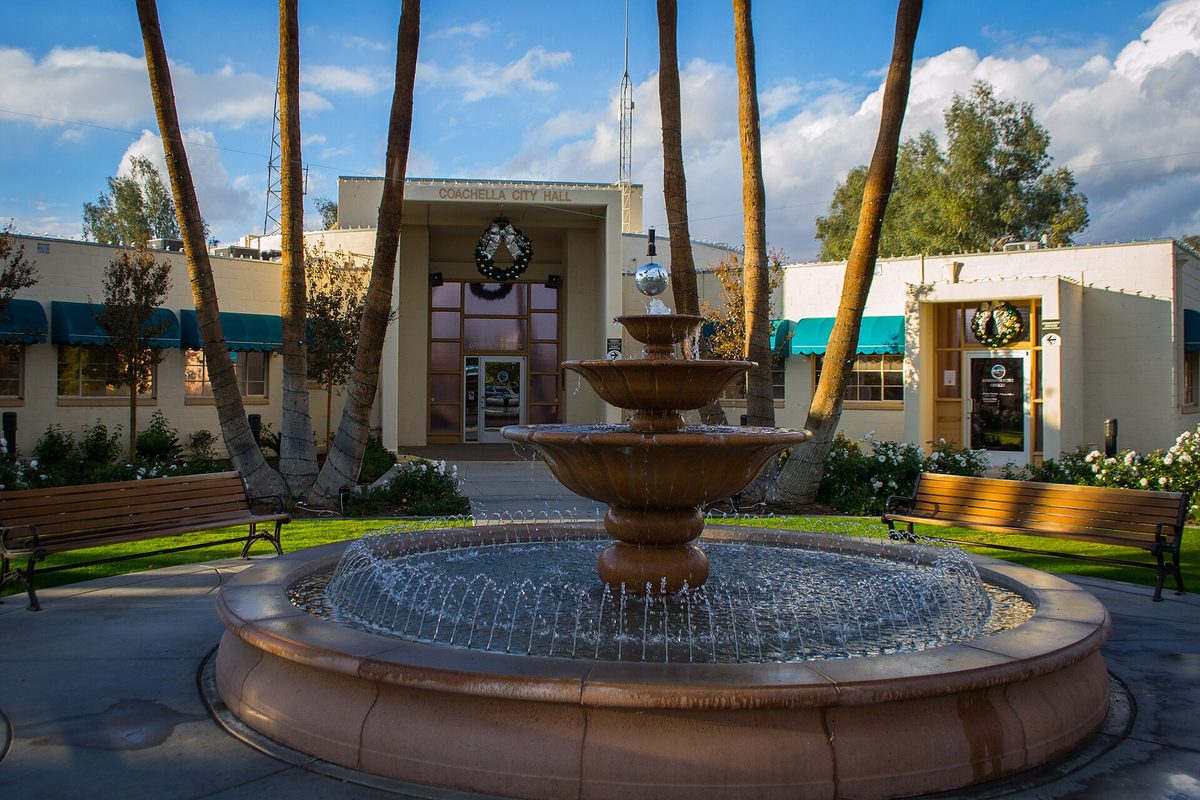 Coachella City Hall behind a tiered stone fountain, framed by tall palm trunks, benches and a green lawn