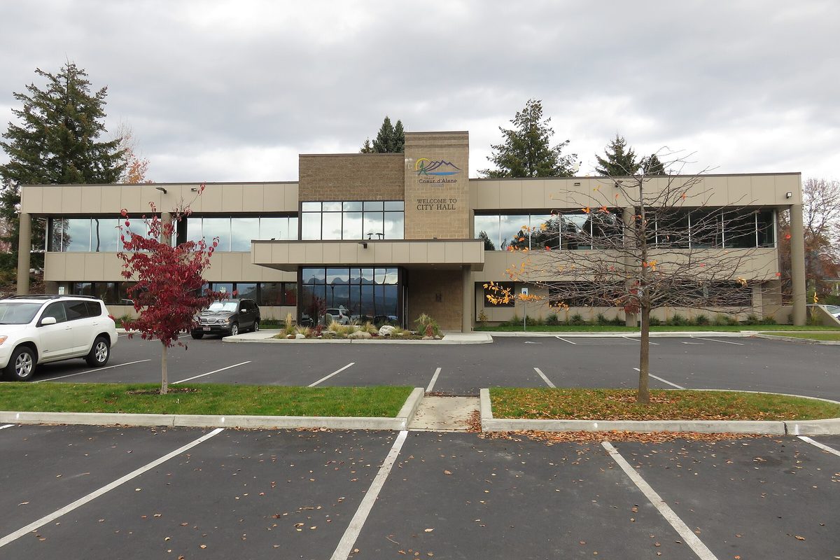 Coeur d'Alene City Hall, a two-story tan building with long bands of windows, seen across a parking lot with autumn trees