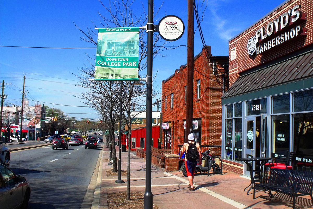 Downtown College Park: a Downtown College Park banner on a lamppost, brick storefronts with a barbershop on the corner, and a man with a backpack walking the sidewalk