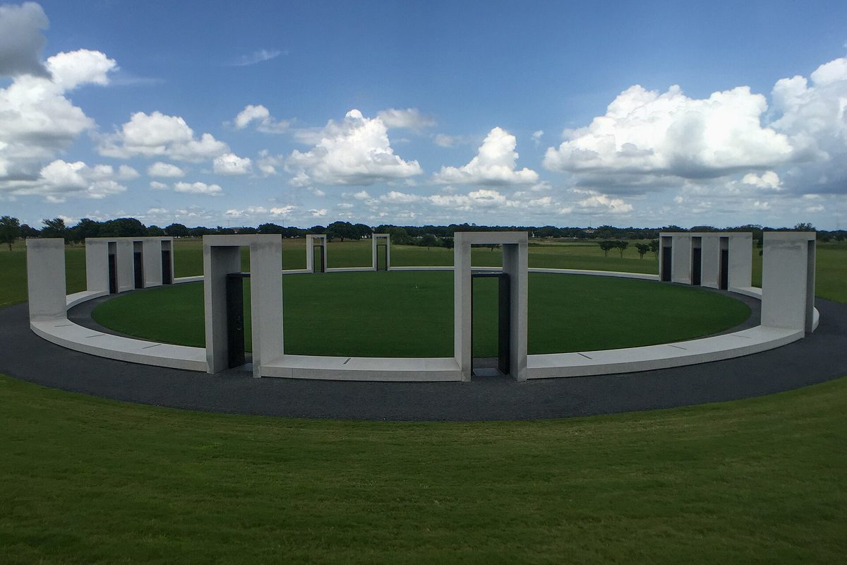 The Bonfire Memorial at Texas A&M: a ring of pale stone portals around a round lawn, open fields and puffy white clouds behind