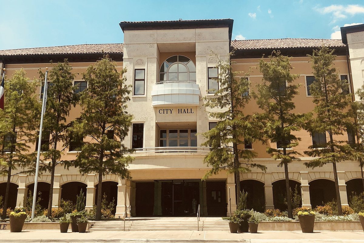 Colleyville City Hall, a tan three-story building with a tile roof, an arched top window and the words City Hall over the entrance, framed by cedar trees