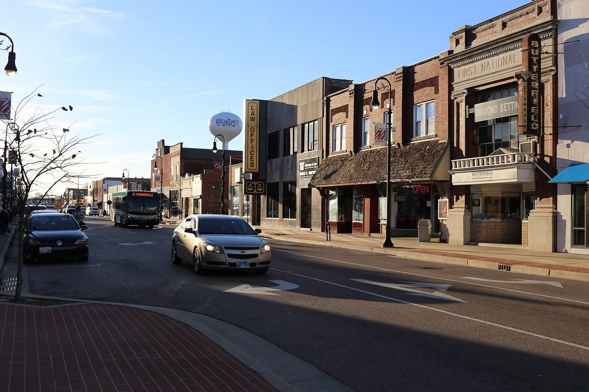Main Street in Collinsville in late-day sun, with brick storefronts, a vertical Law Office sign, a city bus and a car on the road and a water tower behind