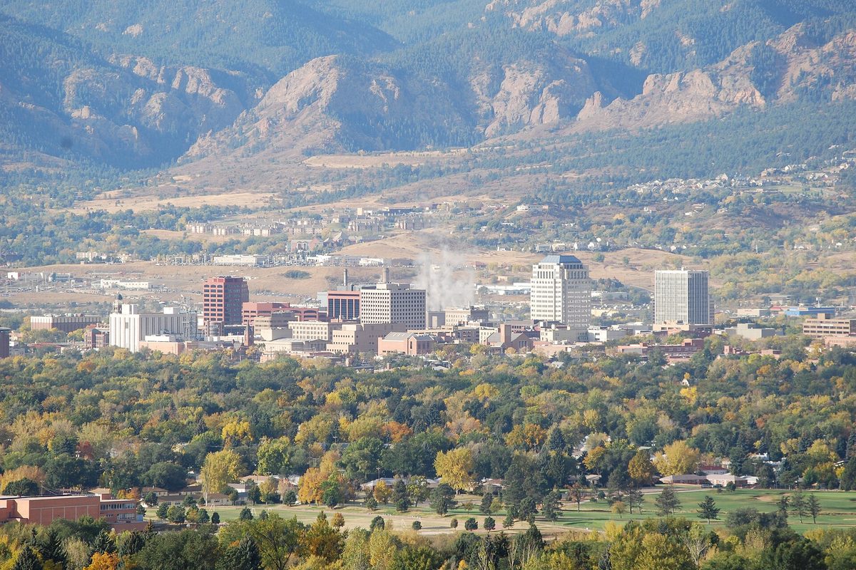 Downtown Colorado Springs office towers rising above autumn trees, with foothills and red rock behind