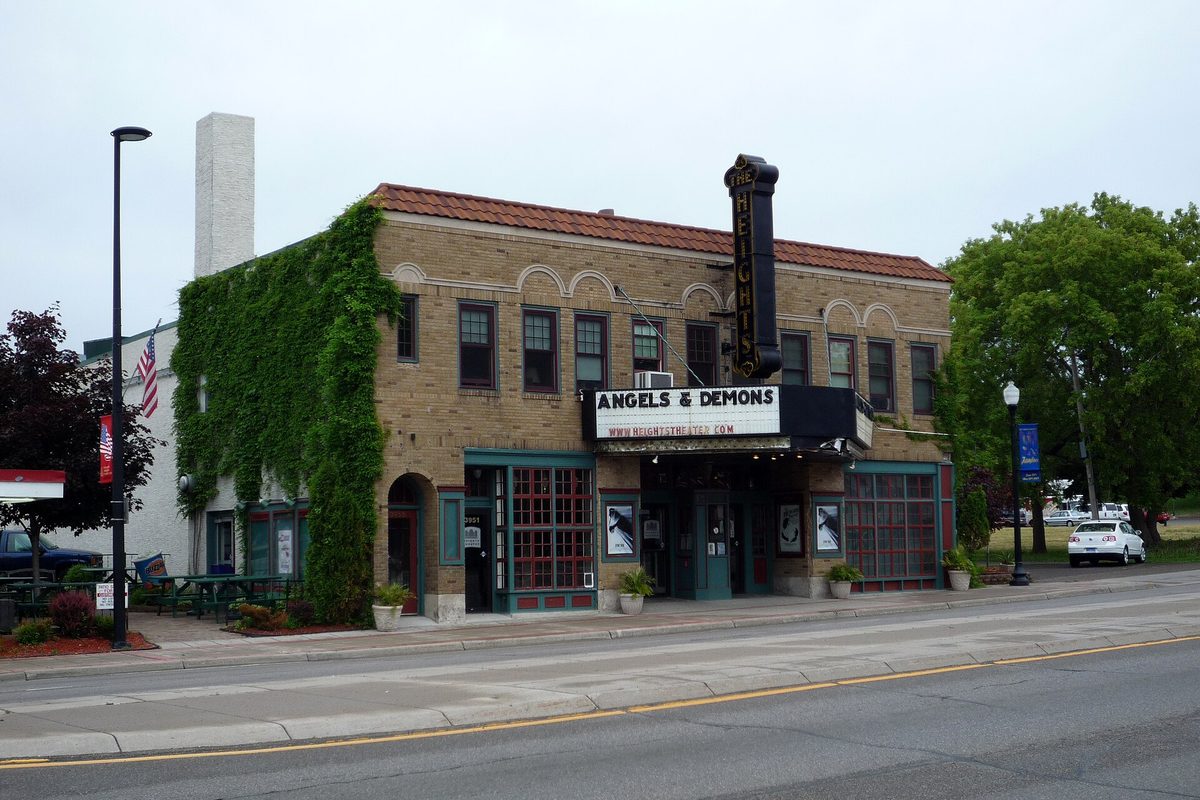 The Heights Theater in Columbia Heights, a tan brick building with a tall vertical sign and an ivy-covered side wall, beside a quiet street under a gray sky