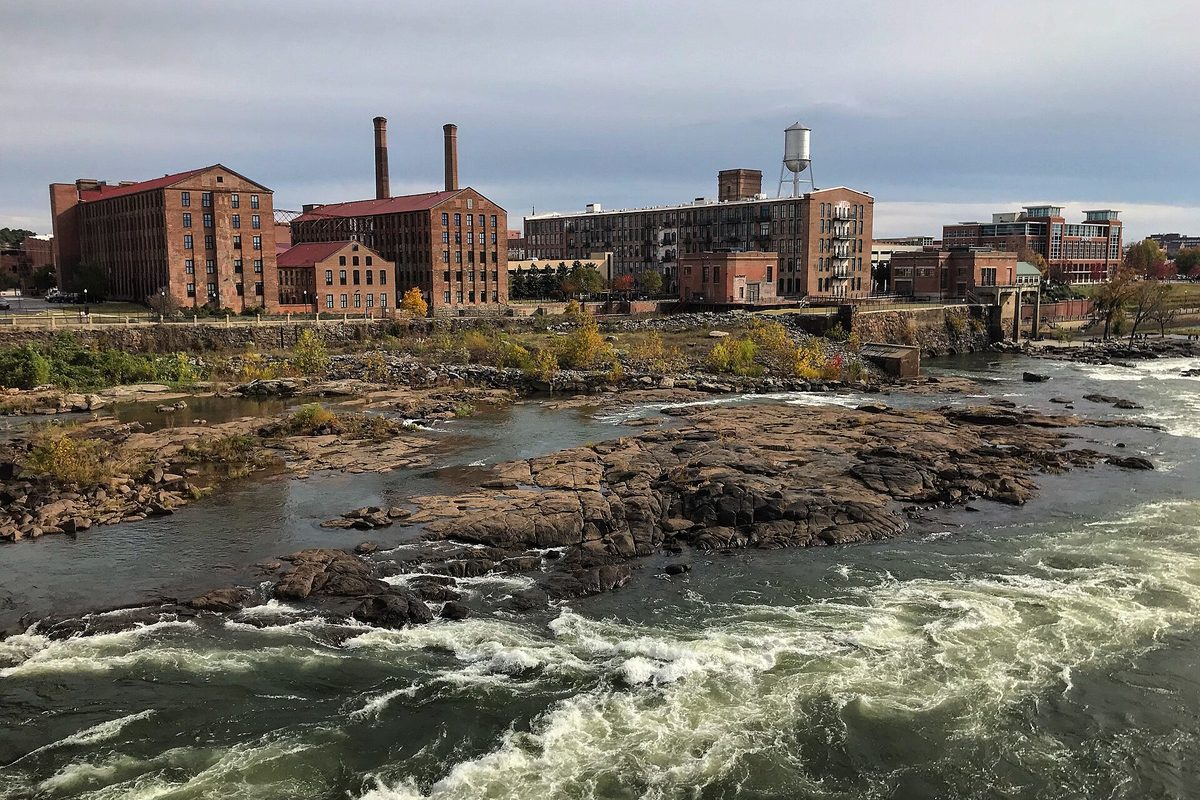 Old red brick mill buildings with tall smokestacks and a water tower above the rocky rapids of the Chattahoochee River in downtown Columbus