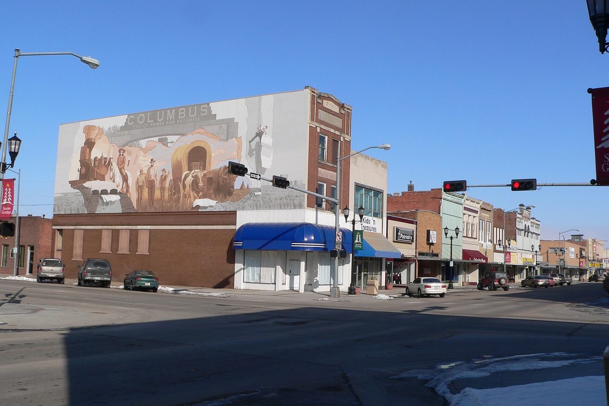 A downtown Columbus corner on 13th Street, with a large mural of covered wagons and pioneers painted on a brick building above blue awnings, on a clear winter day with snow along the curb