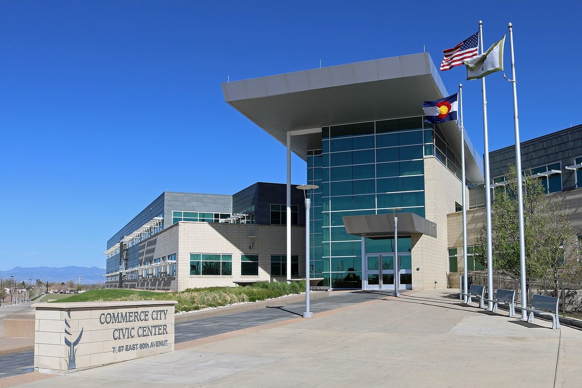 The glass and stone Commerce City Civic Center under a deep blue sky, with American, Colorado and city flags out front and a sign reading 7887 East 60th Avenue