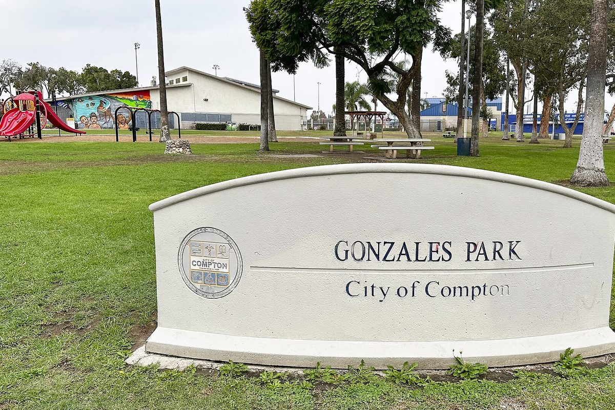The curved white Gonzales Park sign bearing the City of Compton seal, with a red playground slide and a mural-painted building behind it on the lawn