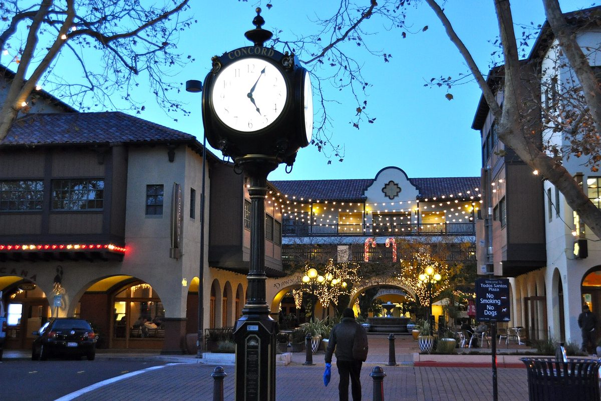 A tall black Concord street clock at Todos Santos Plaza at dusk, with string lights on the arched Spanish-style buildings behind it