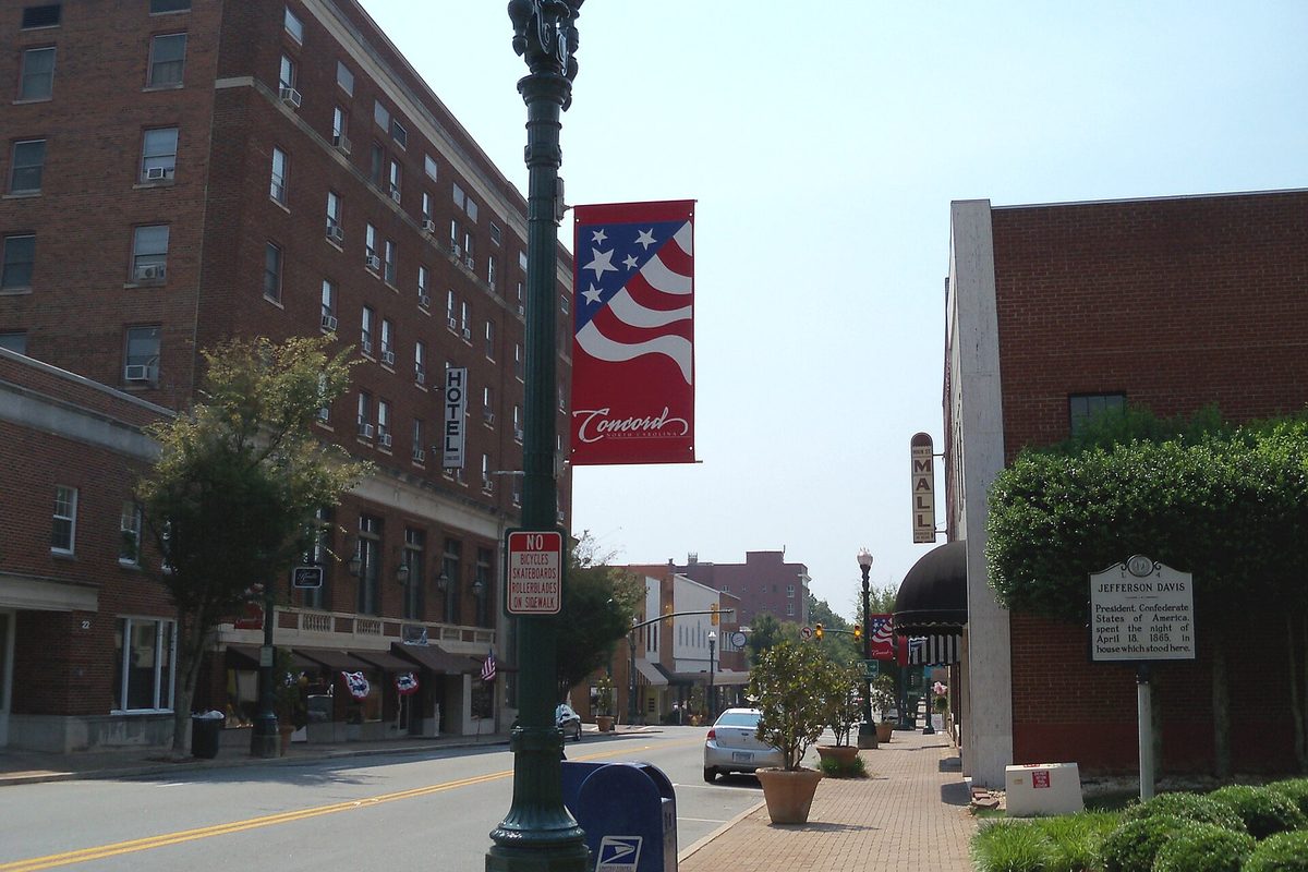 A downtown Concord street with a red, white and blue Concord banner on a green lamppost, a tall brick hotel on the left and a blue mailbox