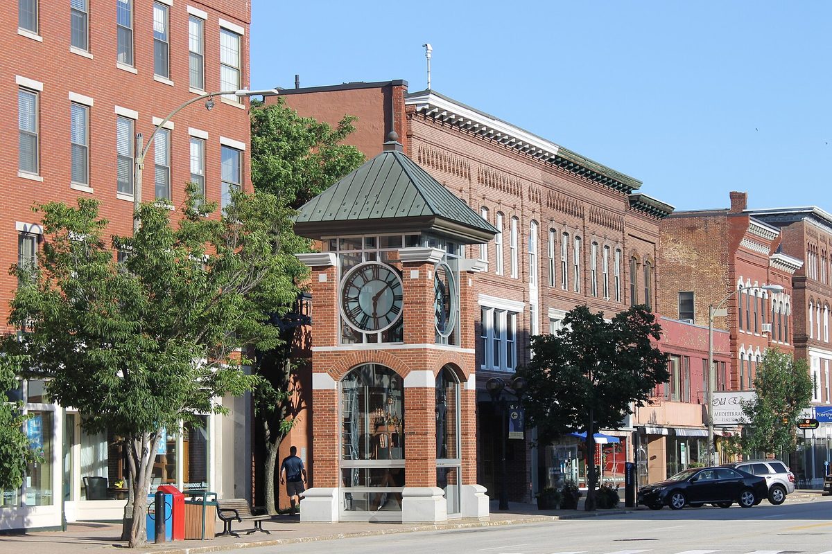 Downtown Concord street with red-brick storefront blocks and a small brick clock tower with a green roof on the sidewalk, trees in leaf and a car parked at the curb