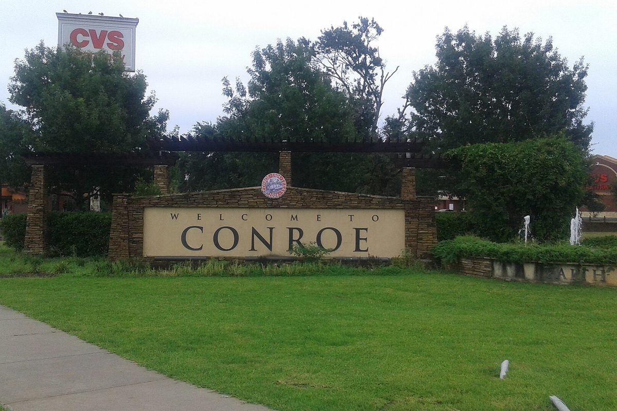 A tan Welcome to Conroe sign set in a stone and brick wall on a green lawn, with trees behind it and a CVS sign rising above the treetops
