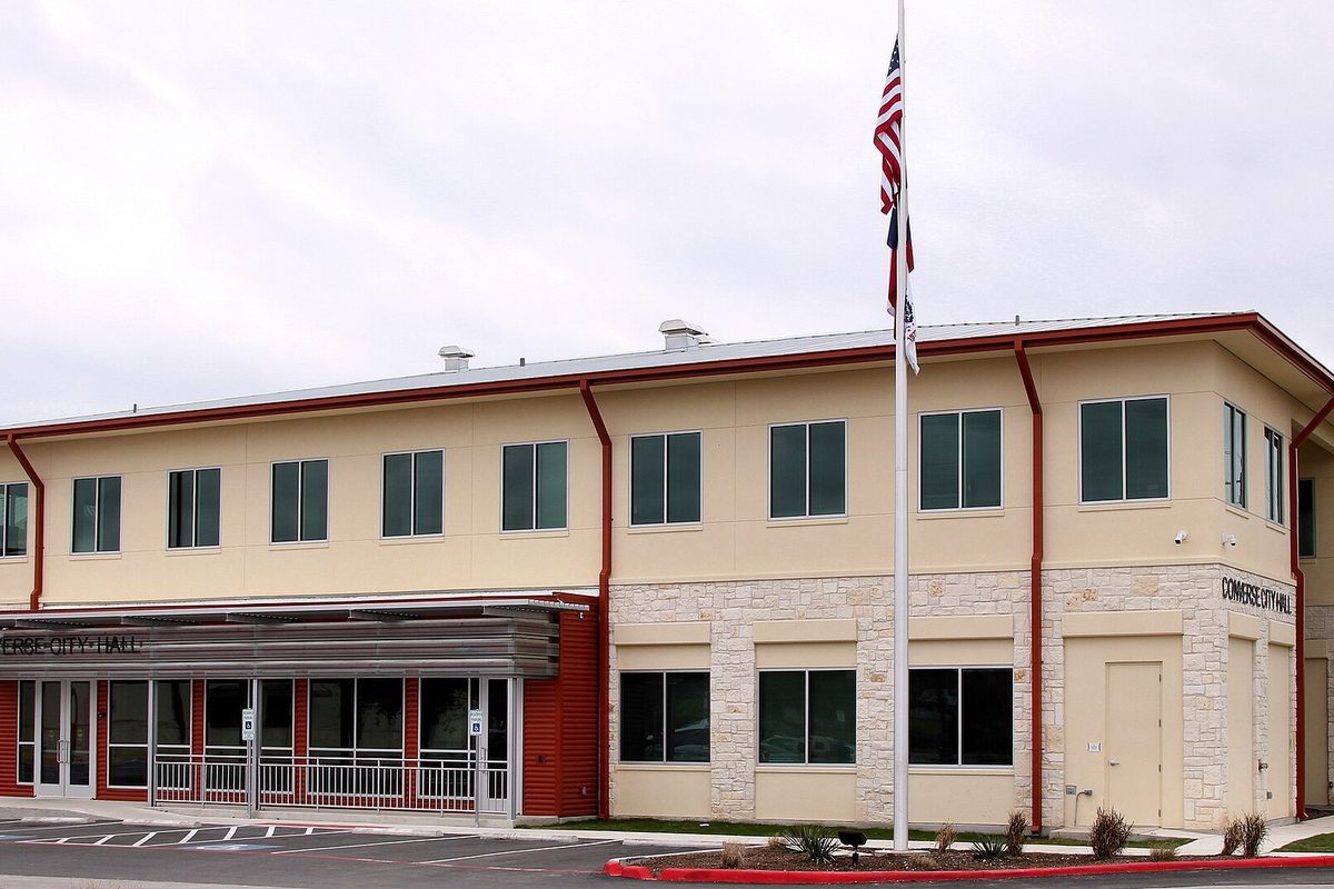 Converse City Hall, a two-story cream building with limestone trim and red downspouts, with an American flag on a pole out front under an overcast sky
