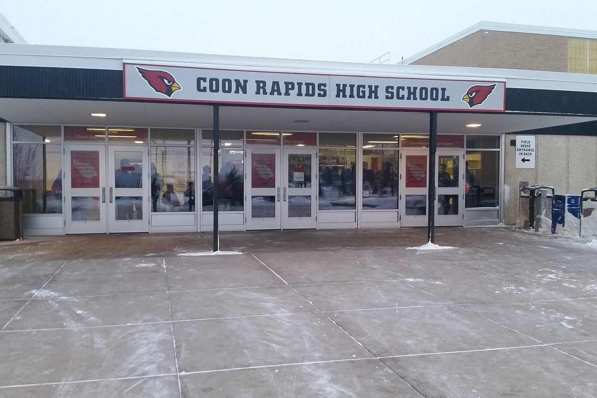 The front entrance of Coon Rapids High School in winter, with a red cardinal sign over the glass doors and a salted walkway
