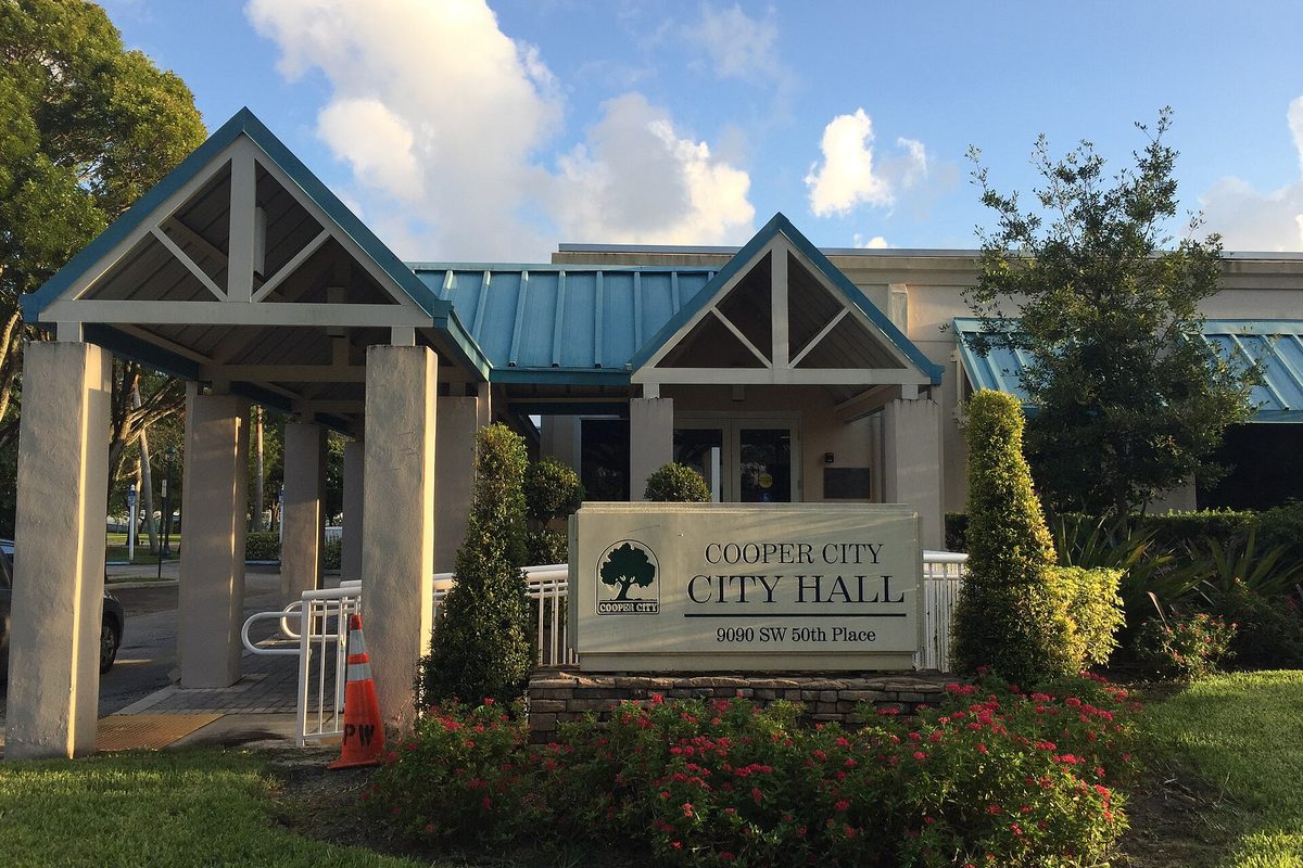 The Cooper City City Hall sign, with a covered entry walkway, red flowers and trees under a blue sky