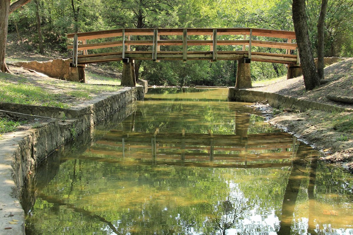 A wooden footbridge on stone piers crossing a still, green stone-lined channel under shade trees in Grapevine Springs Preserve