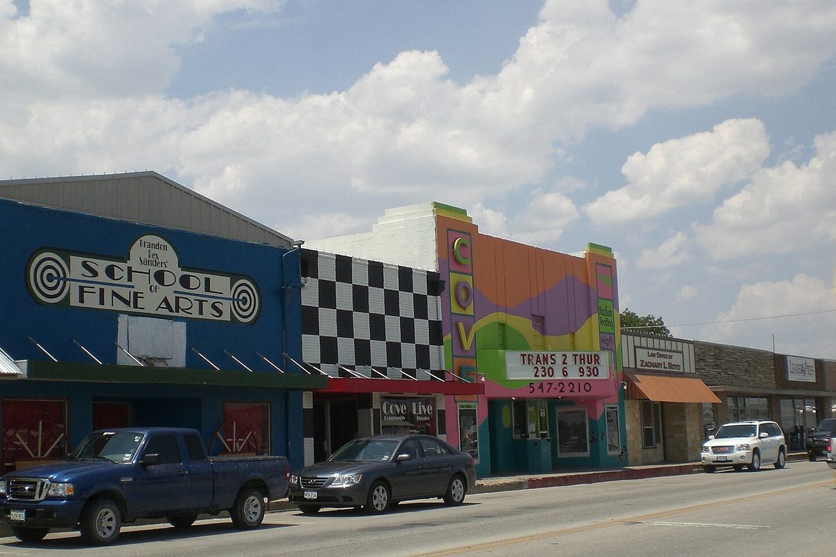 Storefronts on Avenue D in downtown Copperas Cove, with a blue School of Fine Arts building, a checkered facade, a brightly painted theater sign and cars parked at the curb