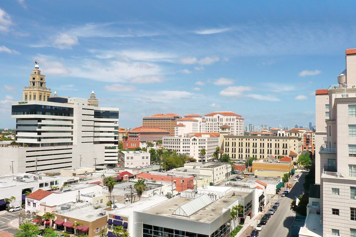 Coral Gables skyline on a clear day: office towers, Mediterranean-style buildings with red tile roofs and a domed tower above low shops and a side street