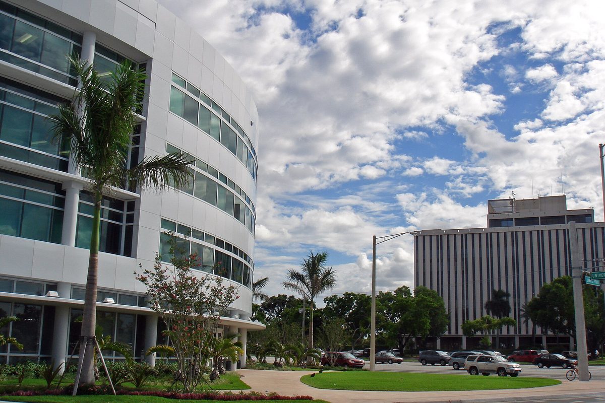 A curved white office building with rows of glass windows and palm trees in front, beside a tall striped office tower and a parking lot in Coral Springs