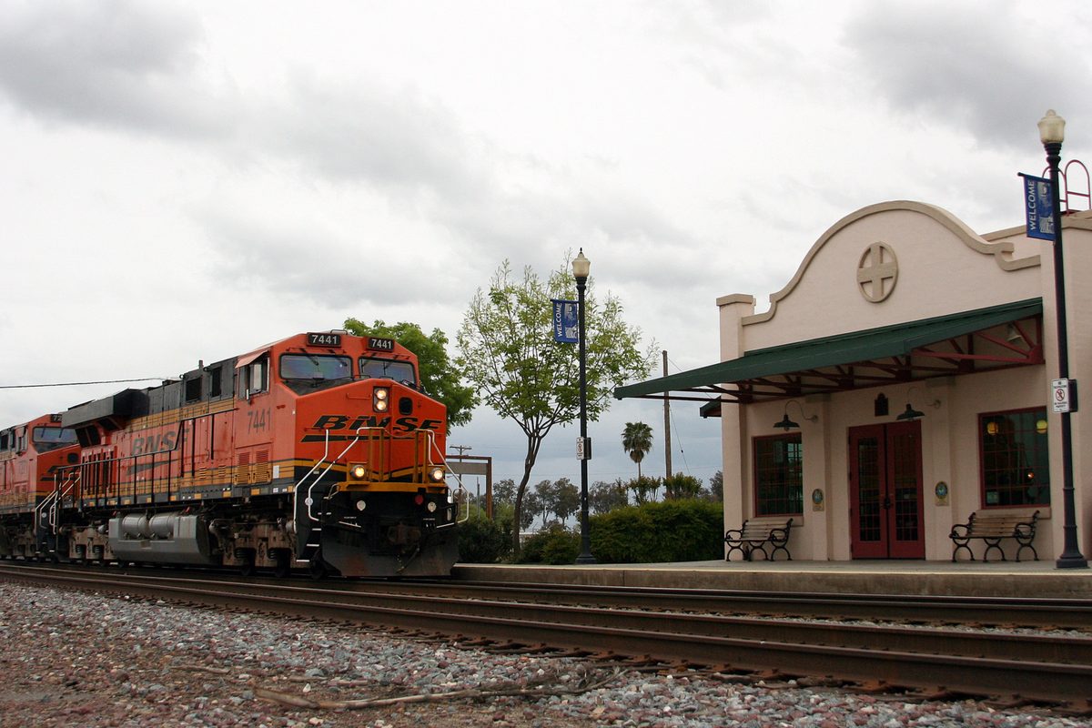 An orange BNSF freight locomotive passing the cream-colored Corcoran Amtrak station with its green awning, benches and lampposts