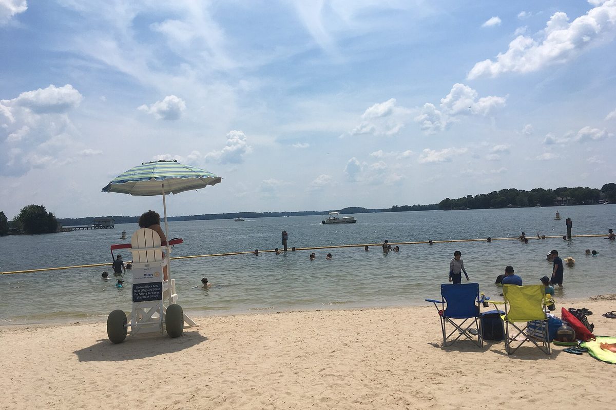A sandy Lake Norman beach in Ramsey Creek Park with a lifeguard chair, a striped umbrella, folding chairs and swimmers inside a rope line