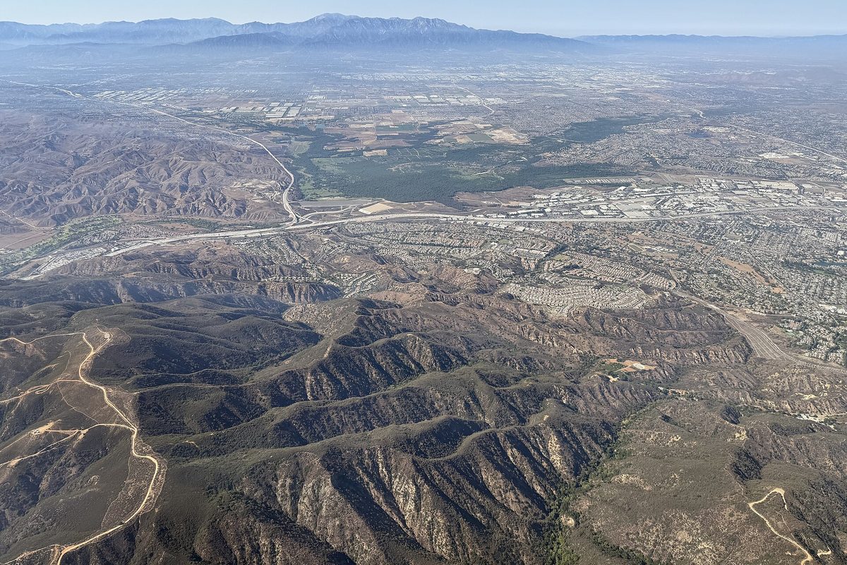 Aerial view of western Corona: brush-covered canyons in front, a freeway and rows of homes beyond, and a mountain range on the hazy horizon