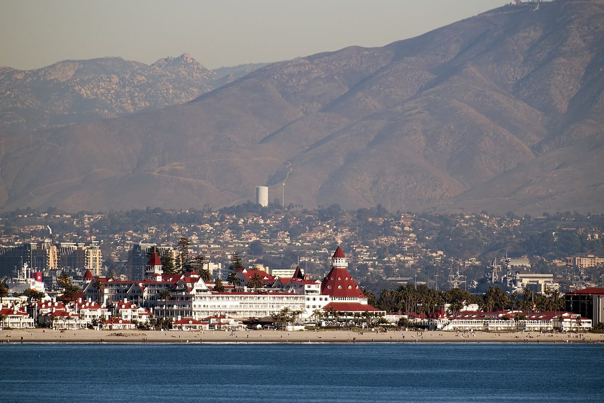 The red-roofed Hotel del Coronado on the beach seen across the water, with hillside neighborhoods and brown mountains behind