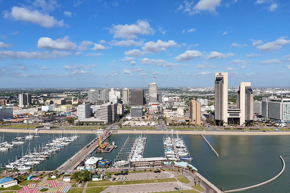 Aerial view of downtown Corpus Christi: towers behind a marina full of sailboats, piers and the seawall, under a wide sky of scattered clouds