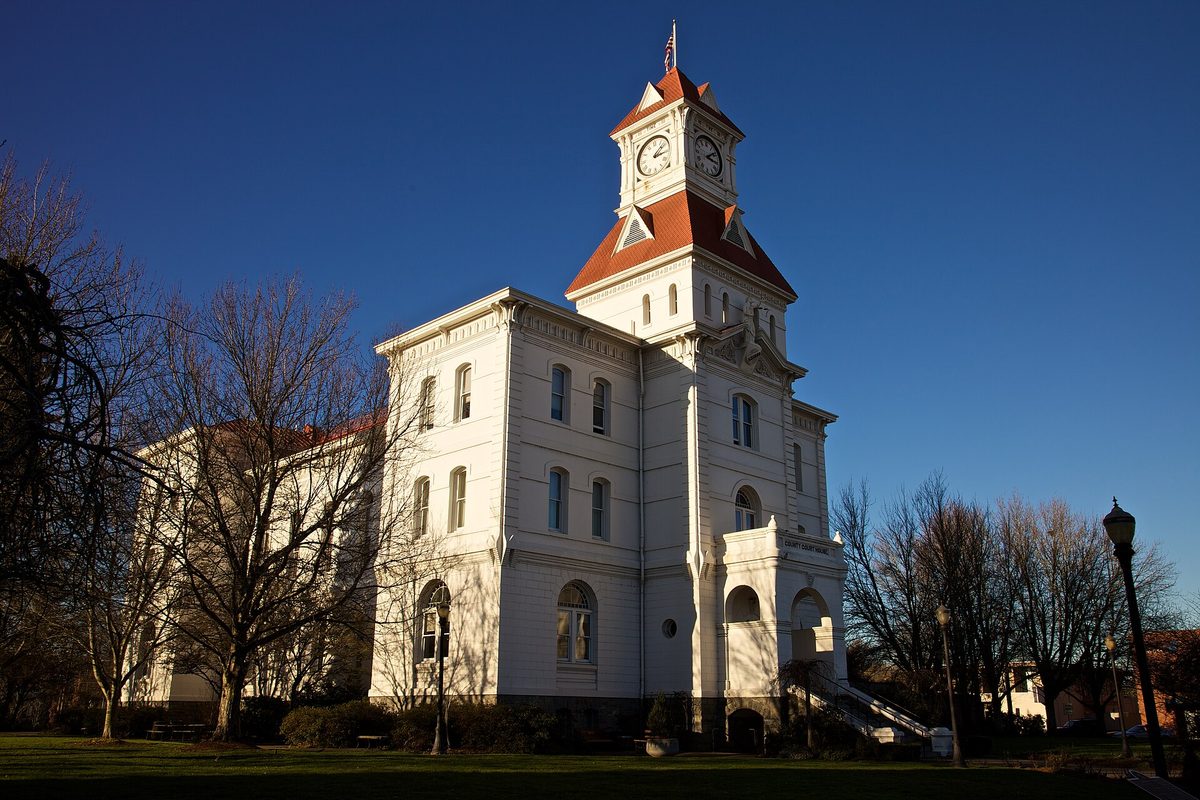 The white Benton County Courthouse in Corvallis, with its red-roofed clock tower against a deep blue sky and bare trees on the lawn
