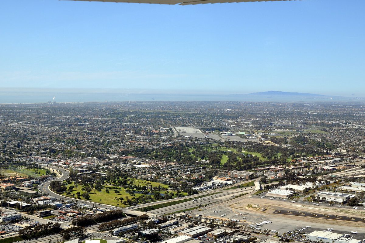 Aerial view over Costa Mesa from a small plane's wing, with a golf course, a freeway, an airport runway and hills in the haze