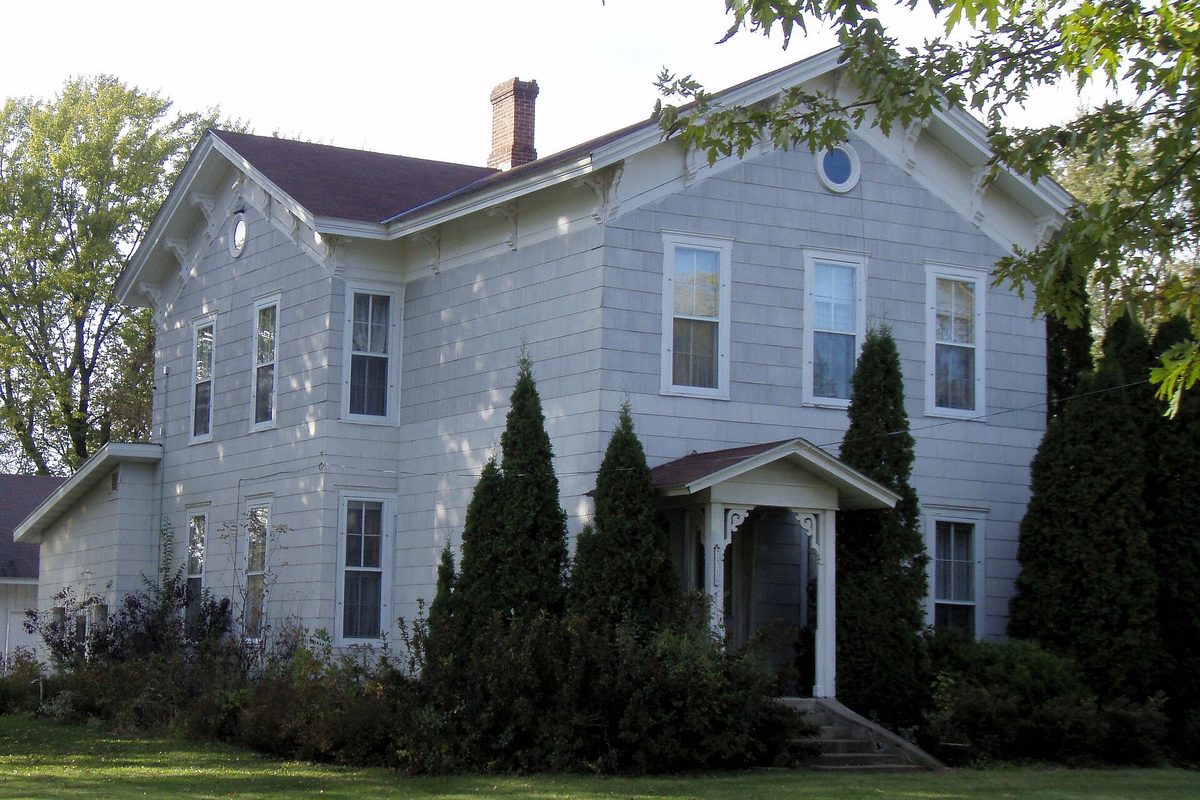 The gray two-story John Furber House in Cottage Grove, with a small front porch and a row of tall evergreens along its front wall