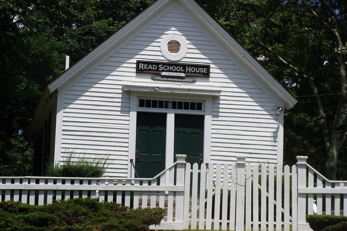 The white clapboard Read School House in Coventry, with green double doors and a round attic vent, behind a white picket fence and dark trees