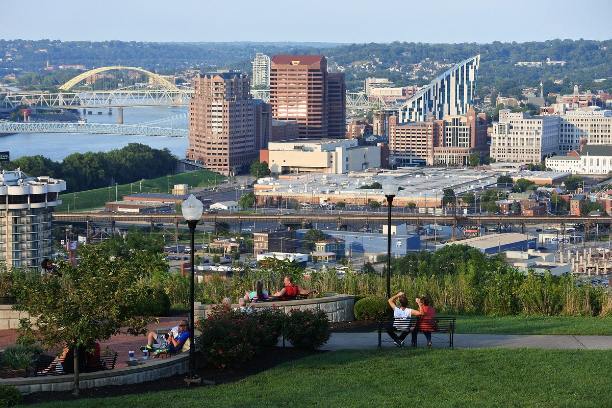 Covington's skyline seen from Devou Park, people on benches in the foreground and the Ohio River with its bridges on the left
