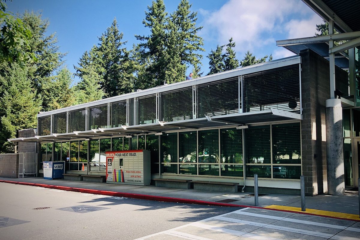 The Covington Library, a long low building with metal sun louvers over its windows, tall evergreens behind it and a book return box by the curb