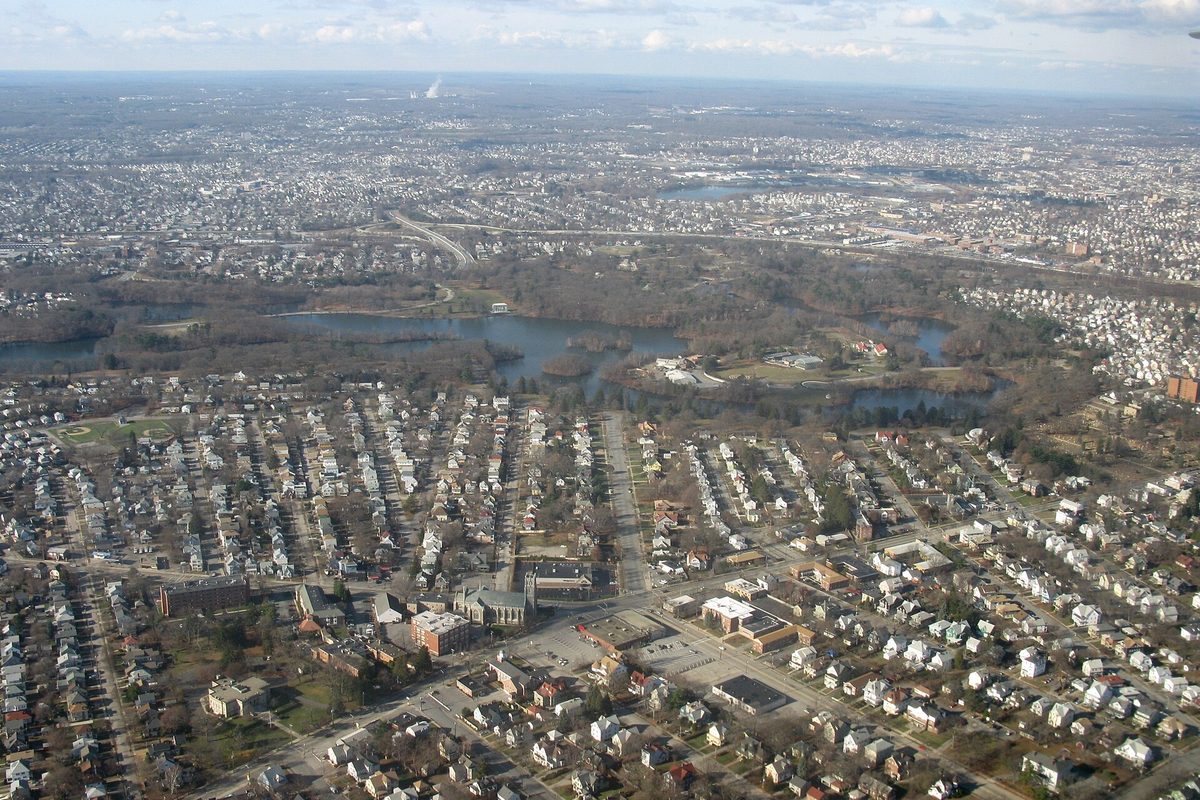 Aerial view over Cranston's rows of houses and streets, with a pond and wooded park in the middle and the wider Providence area stretching to the horizon